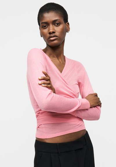 Femme aux cheveux courts portant un haut à manches longues rose style cache-cœur et un pantalon noir, debout les bras croisés devant un fond blanc.