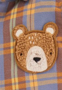 Brauner Teddybär-Patch mit strukturierten Fell-Details, auf blauem, gelbem und orange kariertem Stoffhintergrund aufgenäht.