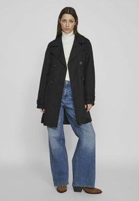 Juodas dvigubas paltas su dideliais mygtukais, plačiomis apykaklėmis ir rankovėmis su atsuktuviu, vilkimas virš baltos golfos ir mėlynų plačių kelnių.