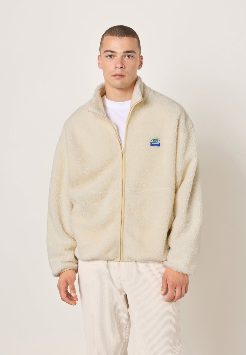 Kermanvärinen fleece-takki, jossa korkea kaulus, täysi vetoketju ja pieni sininen merkki vasemmassa rinnassa. Pehmeä koostumus ja rento istuvuus.