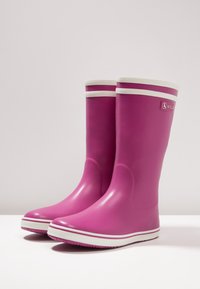 Rosa gummistövlar med vita accenter och sula. Hög design med rundad tå och slät yta. Varumärke på utsidan.