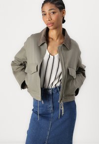Giubbotto bomber verde con colletto in velluto a coste marrone, due tasche frontali, abbinato a una maglietta a righe e una gonna in denim. Tessuto morbido, design corto.
