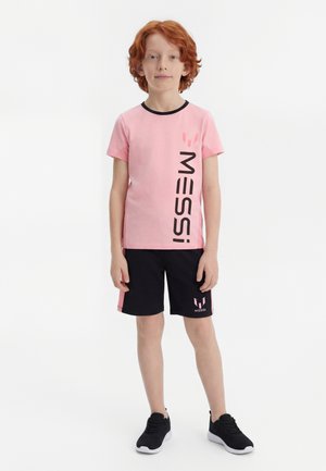 Barn med rødt hår iført en pink Messi T-shirt og sorte shorts, står med armene afslappet mod en hvid baggrund.