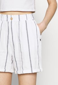 Shorts en lin blanc à rayures verticales noires, taille froncée avec bouton, ourlets retroussés, et une main dans la poche droite.