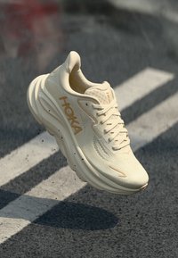 Beige running shoe with textured upper, padded collar, and a thick, cushioned sole. Features HOKA logo in gold on the side.