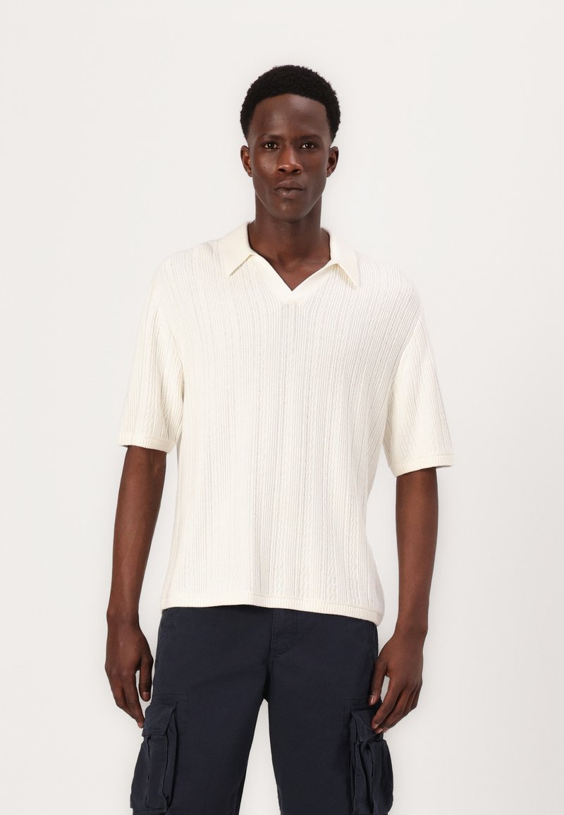 Sweater de malha creme de mangas curtas com decote em V, apresentando textura de riscas verticais e colarinho sutil. Usado com calças cargo escuras.