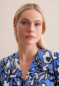 Bluse mit floralem Muster, die einen blauen Hintergrund mit schwarz-weißen Mustern, einen V-Ausschnitt und strukturierter Stoff hat.