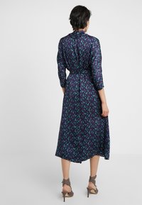 Abito blu navy con motivo a foglie verdi, maniche a tre quarti, vita legata e lunghezza alla caviglia; tessuto morbido con leggero lucido. Tacchi aperti grigi.