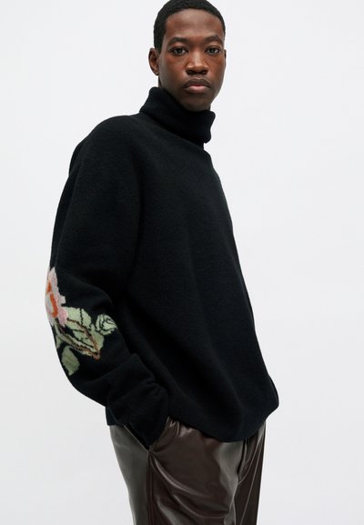 Un tânăr îmbrăcat cu un pulover negru cu guler înalt, având modele florale pe mâneci și pantaloni din piele maro închis, stând în fața unui fundal simplu.