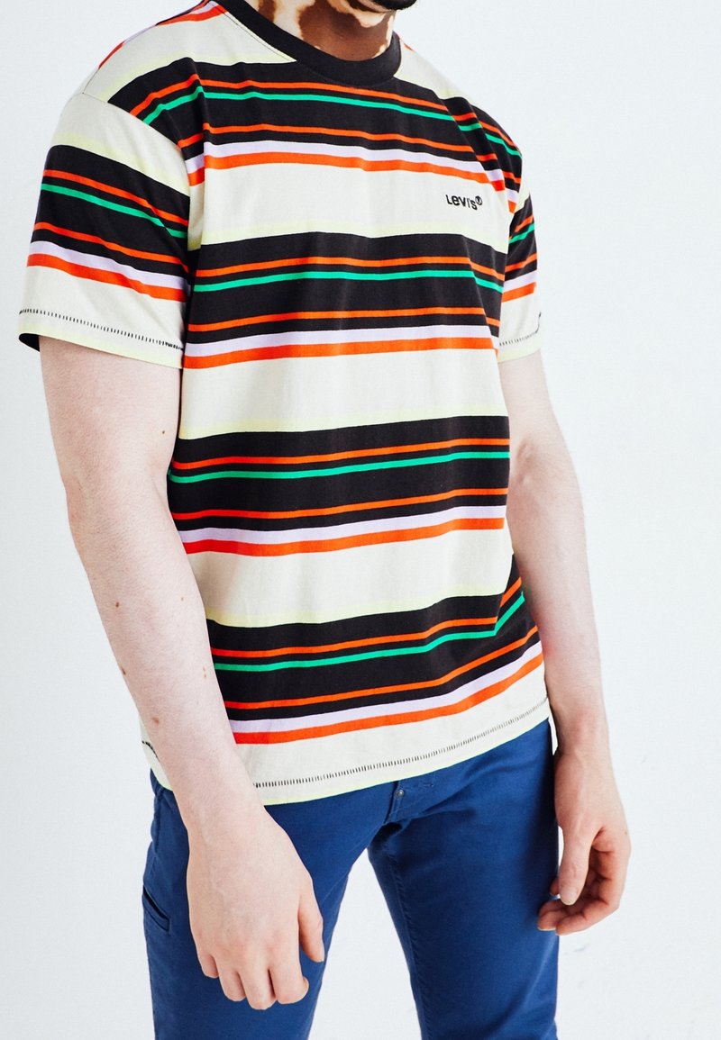 Homme portant un t-shirt Levi's à manches courtes avec des rayures horizontales noires, orange, vertes et blanches, associé à un pantalon bleu.