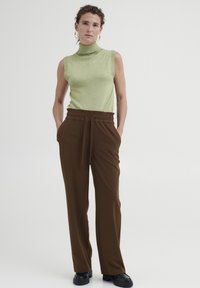 Grüner ärmelloser Rollkragenpullover kombiniert mit braunen hocher tragenden Hosen mit Kordelzug. Schwarze Schuhe vervollständigen den Look.