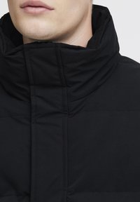 Black padded jacket featuring a high collar, smooth texture, horizontal stitching details, and a minimalist design with no visible hardware.