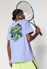 Μωβ βαμβακερό μπλουζάκι με μεγάλο πράσινο και μπλε γραφικό στην πλάτη που απεικονίζει στοιχεία του τένις. Το μοντέλο κρατάει ρακέτα τένις.