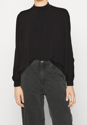 Vrouw die een zwarte blouse met lange mouwen en een hoge hals draagt, gecombineerd met hooggetailleerde donkergrijze spijkerbroeken, staand tegen een effen achtergrond.