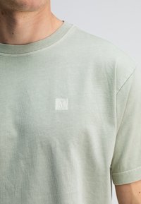 Lichtgroene katoenen t-shirt met ronde hals, korte mouwen en een klein wit logo op de borst. Zacht, gestructureerd materiaal.