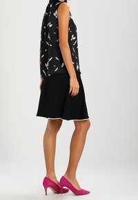 Blouse sans manches noire avec des motifs de feuilles blancs et rouges, associée à une jupe évasée noire et des escarpins en daim rose.