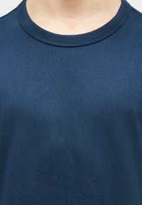 Κοντινό πλάνο ενός ατόμου που φοράει ένα απλό σκουρομπλε πουλόβερ με στρογγυλή λαιμόκοψη, που δείχνει τον λαιμό και την πάνω περιοχή του στήθους.