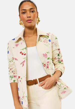 Naine kannab kreemikat jakki mitmevärvilise lillemustriga, valget topi, kreemjaid pükse, pruuni vööd ja kuldseid lehekujulisi kõrvarõngaid.