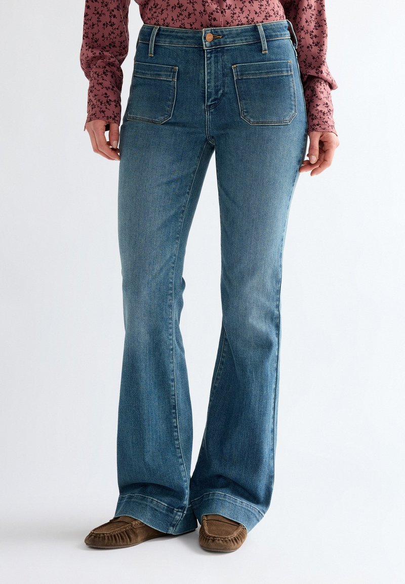 Femme portant un jean bootcut en denim bleu avec des poches plaquées sur le devant, un chemisier rose à fleurs à manches longues et des mocassins marron.