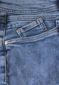 Jeans de mezclilla azul con un detalle en el bolsillo frontal, que presentan costuras a lo largo de los bordes y un cierre de botón metálico. Tejido texturizado con una apariencia desgastada.