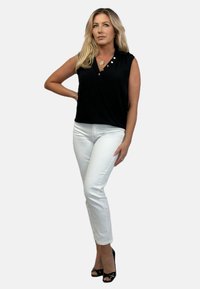 Czarny bezrękawnik o kroju kopertowym, z dwiema guzikami na ramieniu, w połączeniu z białymi spodniami slim fit i czarnymi szpilkami.