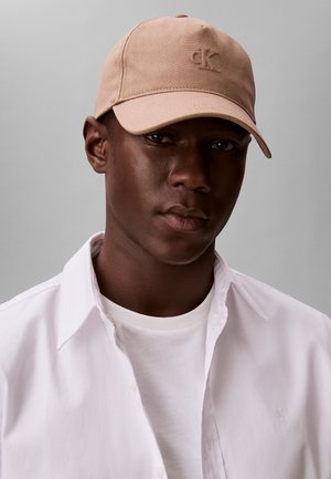 Joven con una gorra beige de Calvin Klein, camisa blanca abotonada sobre una camiseta blanca, sobre un fondo gris liso.