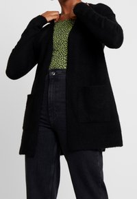 Svart stickad cardigan med lång ärm, öppen front och två framfickor. Bärs över en topp med grönt leopardmönster och svarta högmidjade jeans.