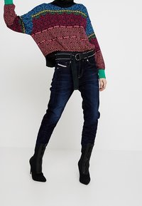 Barevný oversize svetr s multicolor vzory, kombinovaný s tmavými džínami. Obsahuje černý pás a černé vysoké boty.