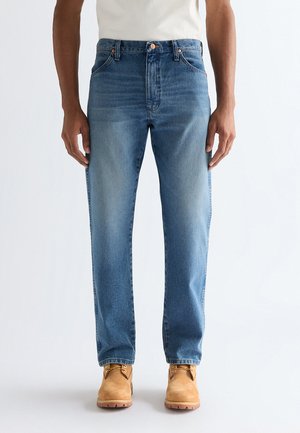 Mann iført lyseblå jeans med rette ben, hvit t-skjorte og beige arbeidssko, stående foran en ensfarget hvit bakgrunn.