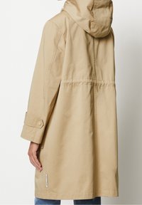 Beige lång kappa med huva och dragsko bak, ärmstraps med knappar, bärs över blå jeans, visad bakifrån i knähöjd.