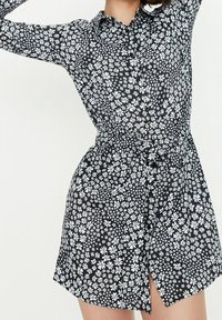 Schwarz geblümtes Hemdkleid mit weißen Blumenmustern, Knopfverschluss, Kragenhöhe und einem Gürtel mit rundem Schnallenverschluss in der Taille.