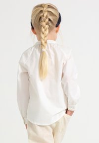 Barn med långt blont hår i fläta, iklädd vit långärmad tröja och ljusa byxor, står med ena handen i fickan och vänder ryggen åt.