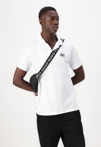 Biely polo tričko s golierom a malým čiernym logom na hrudi, v kombinácii s tmavými nohavicami a čiernou crossbody taškou s bielym brandingom.