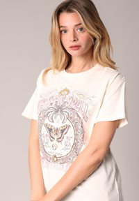 Camiseta de algodón crema con un diseño gráfico colorido que presenta una mariposa y elementos florales, cuello redondo y mangas cortas.