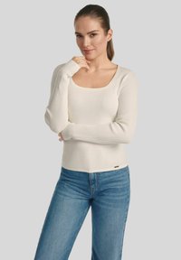 Kobieta z jasnobrązowymi włosami w kucyku, ubrana w dopasowaną, kremową bluzkę z długim rękawem oraz niebieskie dżinsy, stoi z jedną ręką przy brodzie.