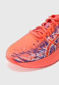 Chaussure de course avec un dessus en mesh corail vif, orné d'un motif dynamique violet, d'une finition texturée et d'une marque blanche contrastante sur le côté.