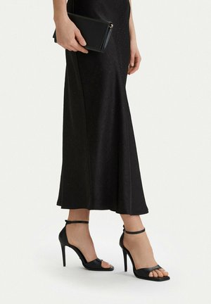 Mujer que lleva tacones altos negros con correa en el tobillo y una falda larga negra, sosteniendo un pequeño bolso de mano negro a su lado.