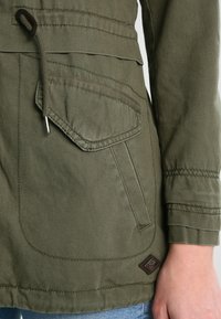 Olivově zelená bunda z bavlněné směsi s nastavovací šňůrkou v pase, velkou přední kapsou a šitými detaily na rukávech. Uvedena hnědá nášivka s logem.