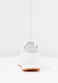 Baskets de sport blanches avec un accent texturé gris au niveau du talon, dotées d'une tige en synthétique, d'un col rembourré et d'une semelle extérieure en caoutchouc marron.