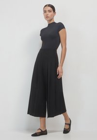 Schwarzes, tailliertes Oberteil mit kurzen Ärmeln, kombiniert mit hochtaillierten, weiten schwarzen Hosen. Das Outfit verfügt über Falten und zarte flache Schuhe.