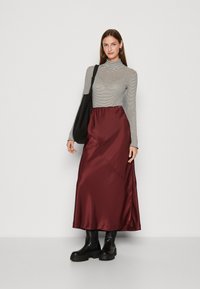 Top cu guler înalt, cu dungi și mâneci lungi, asortat cu o fustă maxi din satin burgund. Modelul poartă cizme negre masive și ține o geantă neagră.