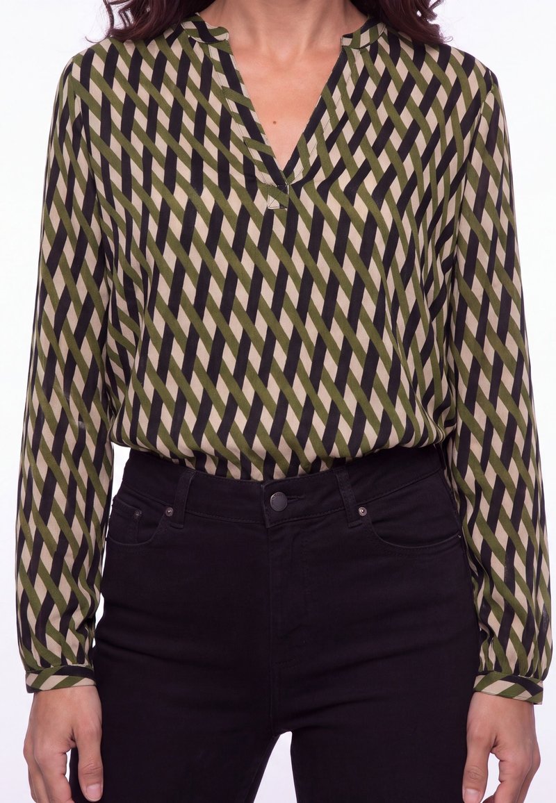 Femme portant un chemisier à manches longues rayé en diagonale vert, beige et noir, rentré dans un pantalon noir taille haute.