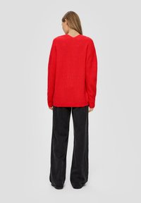 Roter, gerippter Strickpullover mit V-Ausschnitt und überschnittenen Schultern, kombiniert mit weiten schwarzen Hosen. Die Textur wirkt weich und gemütlich.