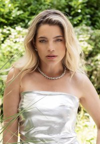 Zilveren strapless jurk met een glanzende textuur, gecombineerd met een sprankelende ketting met ronde, heldere stenen. Groene bladeren op de achtergrond.