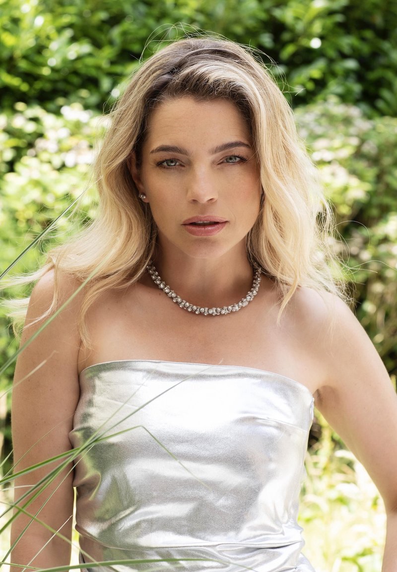 Zilveren strapless jurk met een glanzende textuur, gecombineerd met een sprankelende ketting met ronde, heldere stenen. Groene bladeren op de achtergrond.