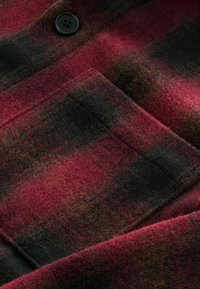 Tissu en laine à carreaux dans des tons de rouge profond, noir et marron. Présente un grand bouton noir et une poche à patch sur une surface texturée.