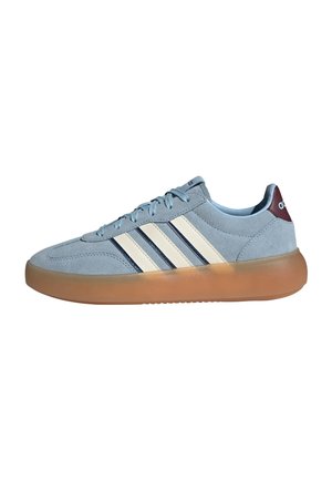 Lichtblauwe suède sneaker met witte en donkerblauwe strepen, bruine doorzichtige zool en rood hieltabje met wit logo.
