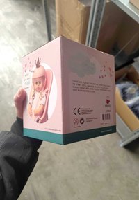 Scatola regalo rosa con una bambola con una corona. Contiene grafiche giocose, loghi di sicurezza e testo. La base è turchese, con un'etichetta non tossica.