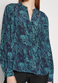 Blouse marine avec un motif marbré vert, dotée d'un col en V et de manches longues. Le tissu semble lisse avec une finition légèrement brillante.