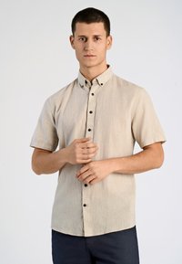 Camisa de botones de manga corta en color beige, confeccionada en una tela texturizada. Presenta un cuello puntiagudo y botones oscuros, combinada con pantalones oscuros.